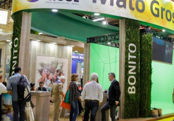 Trade show booth for Mato Grosso do Sul at a convention; visitors gather near a reception desk under a curved banner that reads 'Mato Grosso do Sul', with green vertical columns labeled 'Bonito' surrounding the space.
