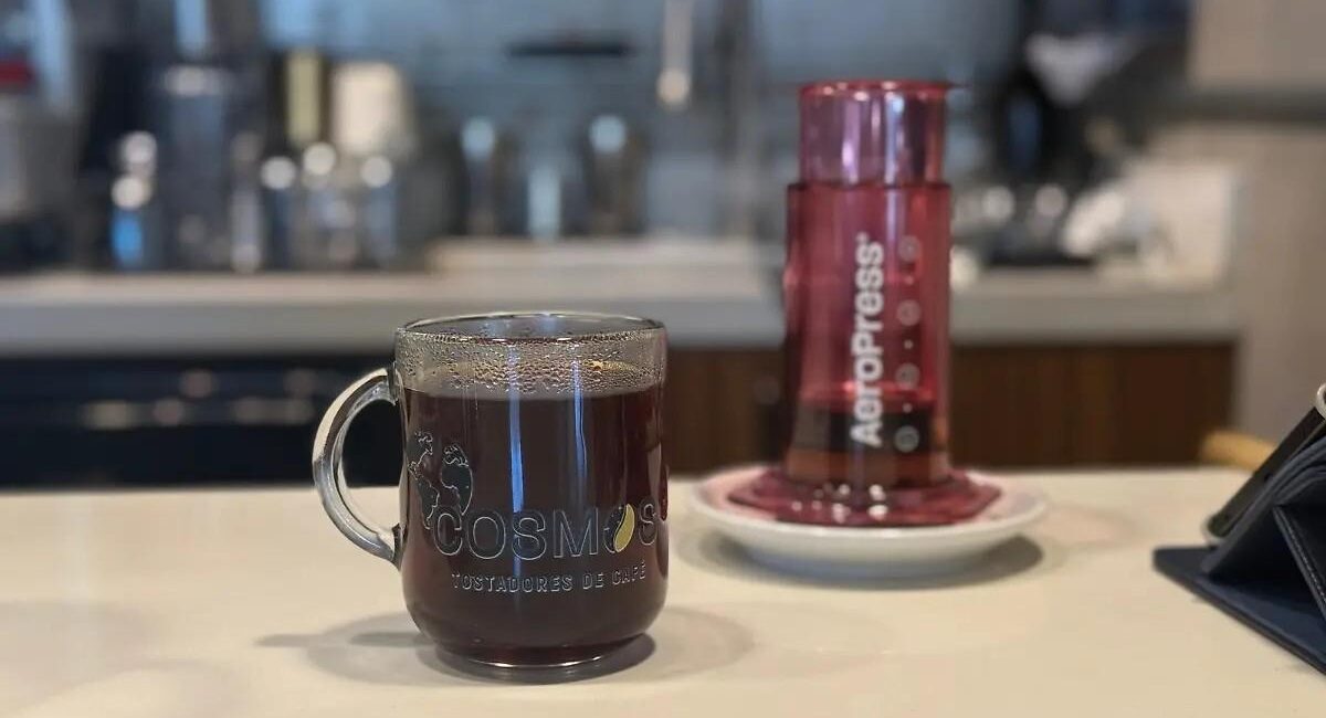 Glass mug of coffee labeled COSMOS on a cafe counter, with a pink Aeropress device in the background.