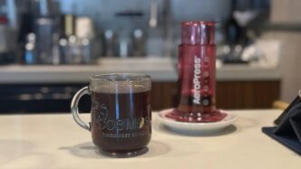 Glass mug of coffee labeled COSMOS on a cafe counter, with a pink Aeropress device in the background.