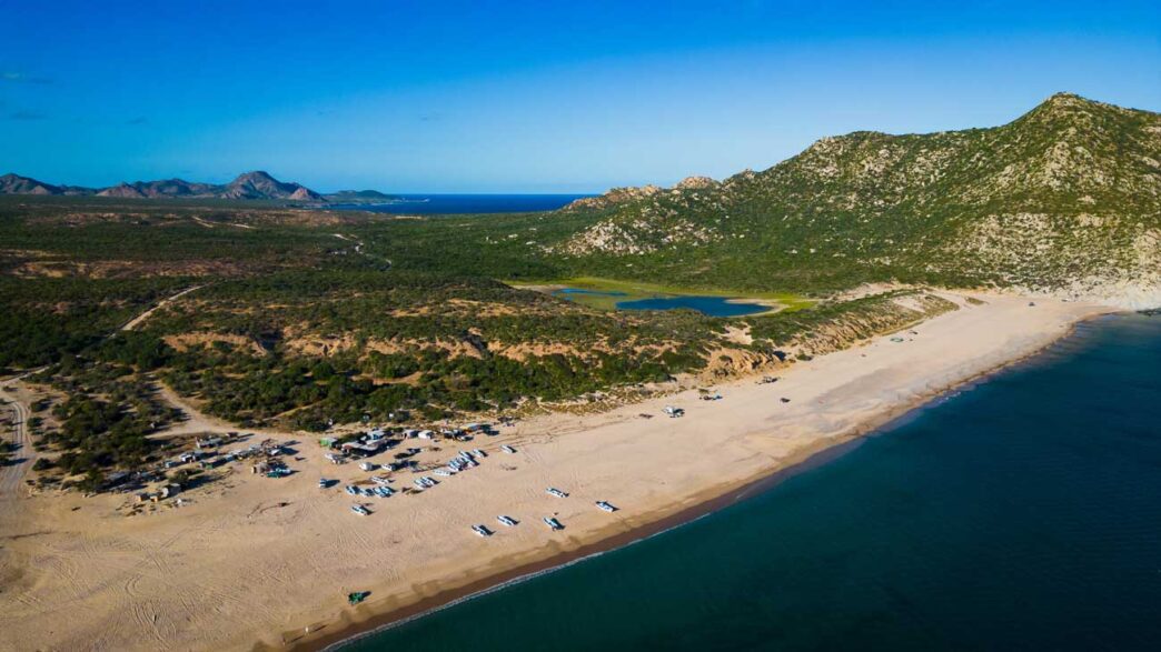 caboPulmo - Gringo Gazette Aerial view of a sandy beach with many small boats lined up along the shore, turquoise water, and green hills in the distance with rocky mountains beyond.
