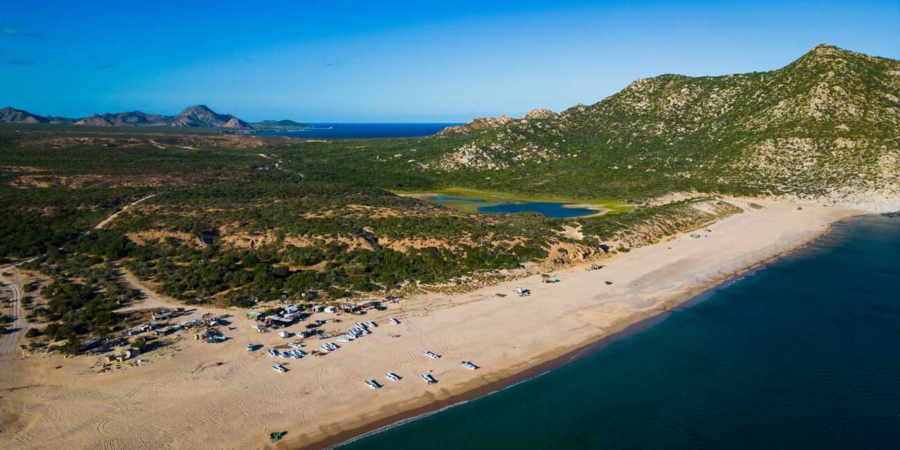 Aerial view of a sandy beach with many small boats lined up along the shore, turquoise water, and green hills in the distance with rocky mountains beyond.