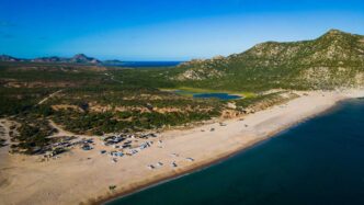 Aerial view of a sandy beach with many small boats lined up along the shore, turquoise water, and green hills in the distance with rocky mountains beyond.