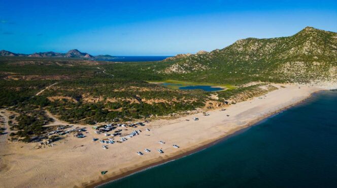 Aerial view of a sandy beach with many small boats lined up along the shore, turquoise water, and green hills in the distance with rocky mountains beyond.