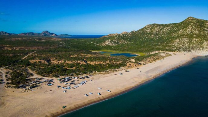 Aerial view of a sandy beach with many small boats lined up along the shore, turquoise water, and green hills in the distance with rocky mountains beyond.