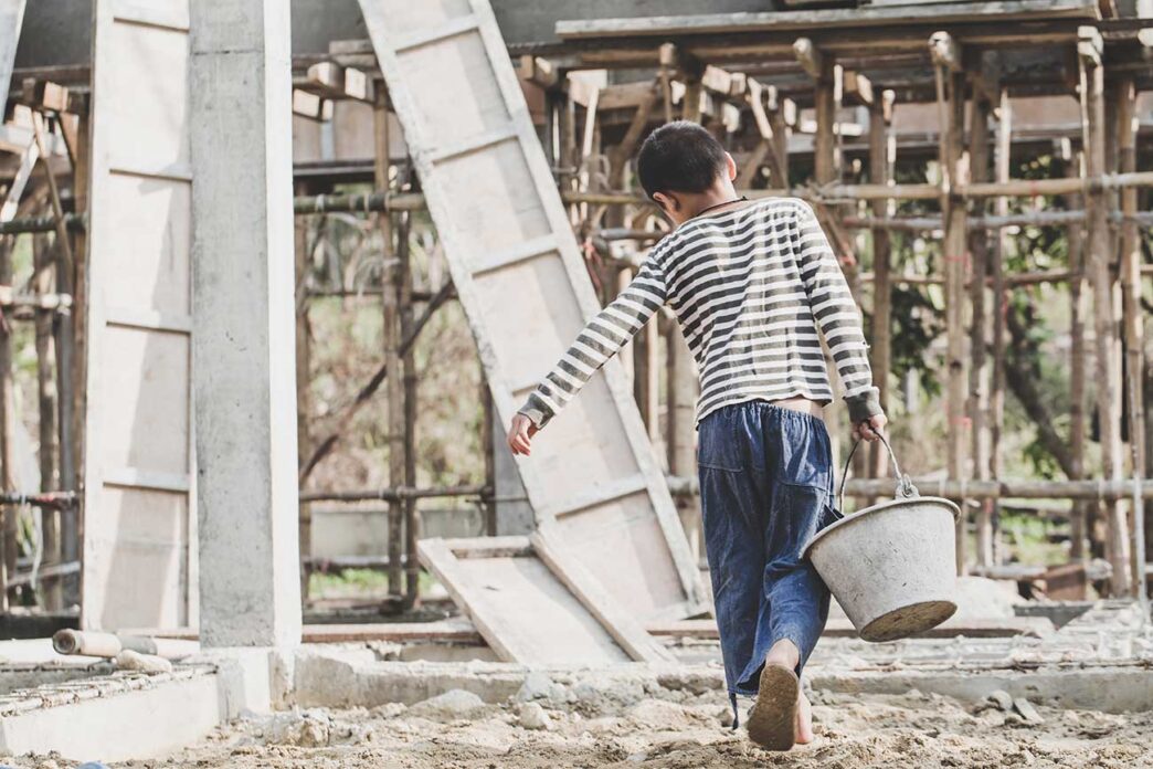childLabor - Gringo Gazette Person in striped shirt and blue pants walks away barefoot on a dusty construction site, carrying a metal bucket with scaffolding in the background.