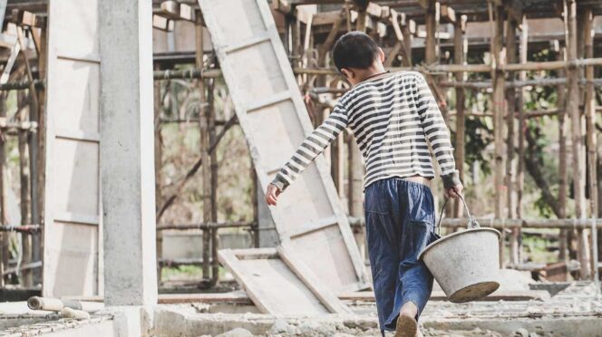 Person in striped shirt and blue pants walks away barefoot on a dusty construction site, carrying a metal bucket with scaffolding in the background.