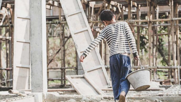 Person in striped shirt and blue pants walks away barefoot on a dusty construction site, carrying a metal bucket with scaffolding in the background.