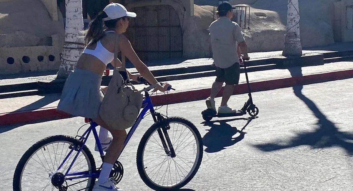 Woman wearing a white cap rides a blue bicycle on a sunny street, carrying a beige bag on the frame.