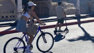 Woman wearing a white cap rides a blue bicycle on a sunny street, carrying a beige bag on the frame.