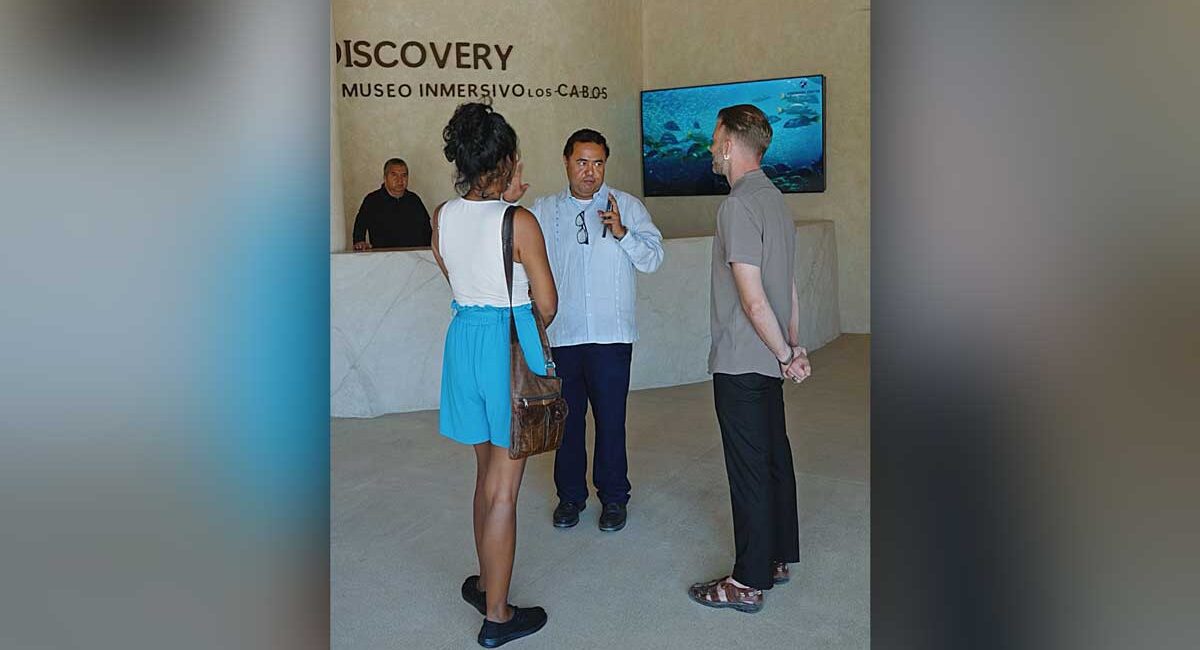 Group of three adults conversing in a museum lobby, with a marble reception desk and an underwater video on a screen in the background.
