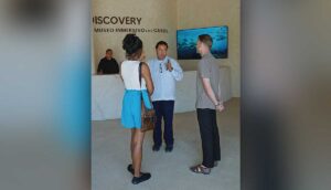 Group of three adults conversing in a museum lobby, with a marble reception desk and an underwater video on a screen in the background.