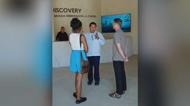 Group of three adults conversing in a museum lobby, with a marble reception desk and an underwater video on a screen in the background.