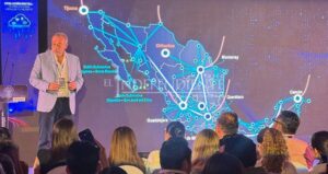 Presenter on stage beside a large screen displaying a blue, connected map of Mexico with cities and routes; audience seated in front of the stage at a conference.