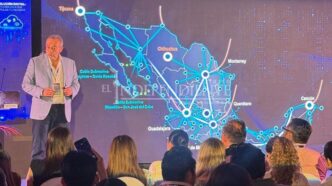 Presenter on stage beside a large screen displaying a blue, connected map of Mexico with cities and routes; audience seated in front of the stage at a conference.