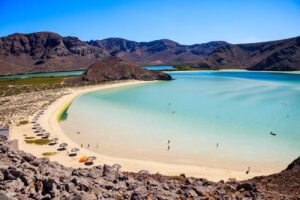 Turquoise lagoon with white sandy beach and rows of umbrellas, encircled by rocky hills under a clear blue sky.