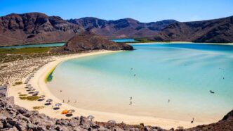 Turquoise lagoon with white sandy beach and rows of umbrellas, encircled by rocky hills under a clear blue sky.