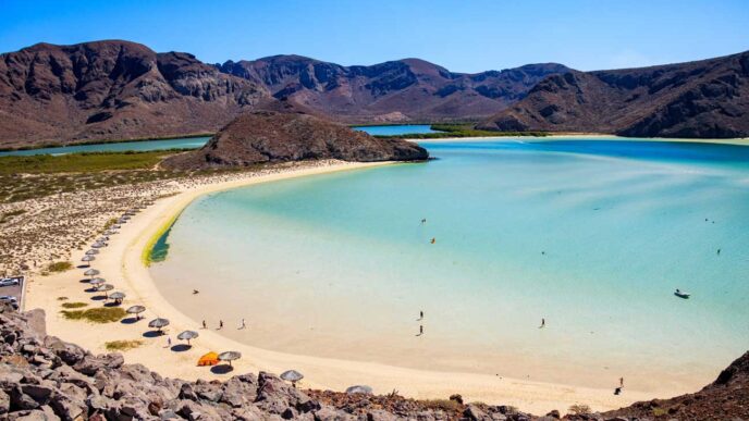 Turquoise lagoon with white sandy beach and rows of umbrellas, encircled by rocky hills under a clear blue sky.