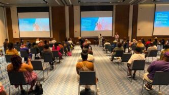Conference room full of attendees facing a stage with speakers and large projection screens.