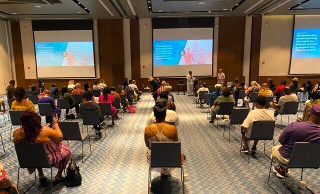 Conference room full of attendees facing a stage with speakers and large projection screens.