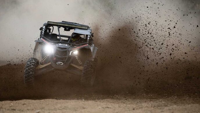 Off-road UTV kicking up a large cloud of dirt on a dusty track, headlights on.