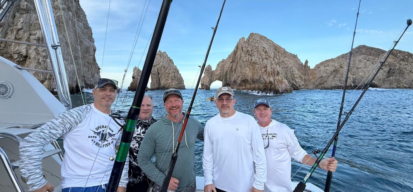 Five men on a fishing boat posing with rods, rocky coastline and an arch in the background under a clear blue sky