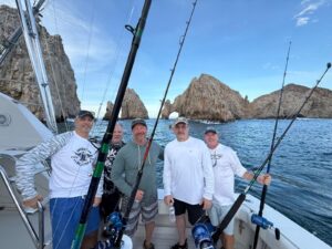 Five men on a fishing boat posing with rods, rocky coastline and an arch in the background under a clear blue sky