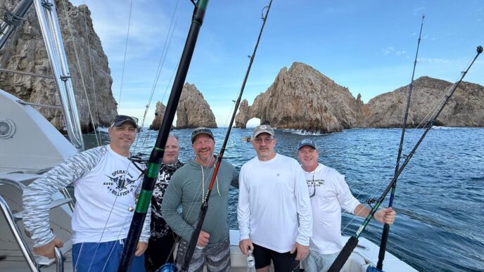 Five men on a fishing boat posing with rods, rocky coastline and an arch in the background under a clear blue sky