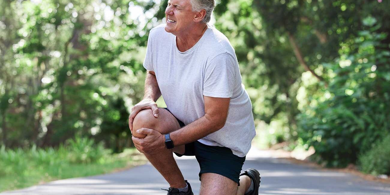 Older man in a gray t‑shirt and black shorts kneels on a park path, stretching his knee outdoors in sunlight.