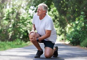 Older man in a gray t‑shirt and black shorts kneels on a park path, stretching his knee outdoors in sunlight.