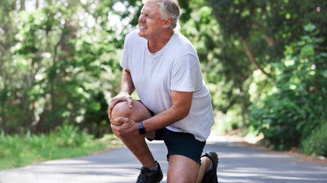 Older man in a gray t‑shirt and black shorts kneels on a park path, stretching his knee outdoors in sunlight.