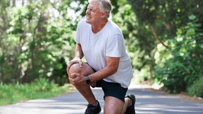 Older man in a gray t‑shirt and black shorts kneels on a park path, stretching his knee outdoors in sunlight.