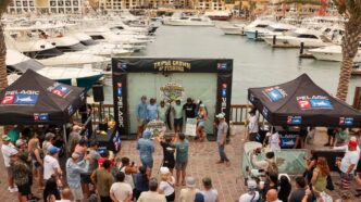 Crowd gathered at a marina festival around a stage labeled 'Triple Crown of Fishing' with boats docked in the background under tents labeled Pelagic P.