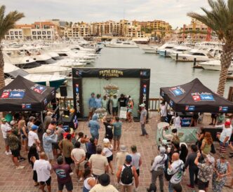 Crowd gathered at a marina festival around a stage labeled 'Triple Crown of Fishing' with boats docked in the background under tents labeled Pelagic P.