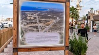 Wood-framed informational map board on a seaside promenade, showing a coastal desert area with roads and a blue sea in the distance.