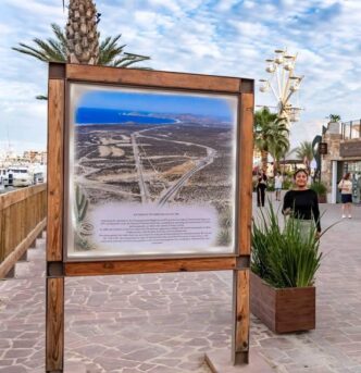 Wood-framed informational map board on a seaside promenade, showing a coastal desert area with roads and a blue sea in the distance.