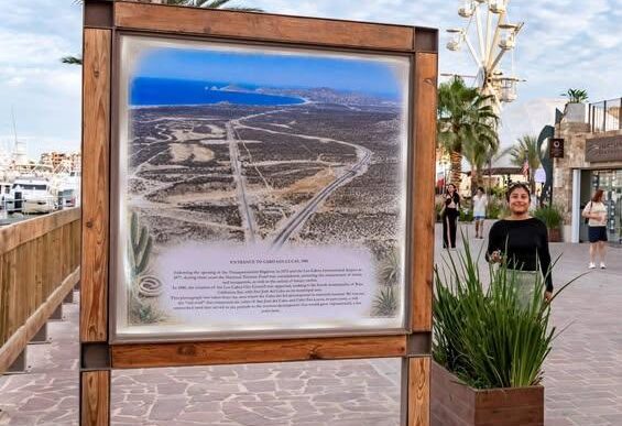 Wood-framed informational map board on a seaside promenade, showing a coastal desert area with roads and a blue sea in the distance.