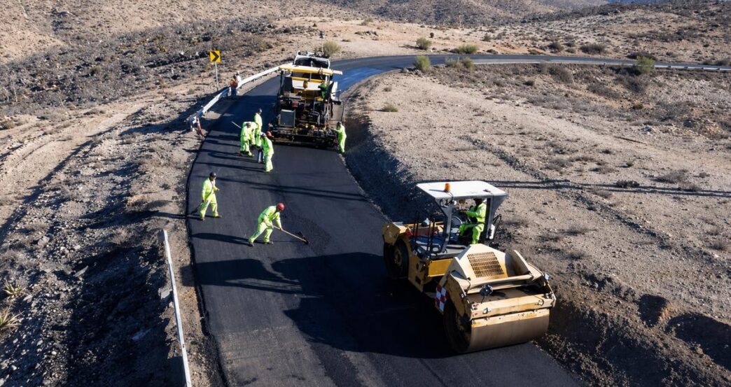 Road construction crew in high-visibility outfits laying asphalt on a winding desert road with a roller and paving machine nearby.