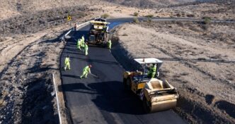 Road construction crew in high-visibility outfits laying asphalt on a winding desert road with a roller and paving machine nearby.
