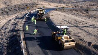 Road construction crew in high-visibility outfits laying asphalt on a winding desert road with a roller and paving machine nearby.