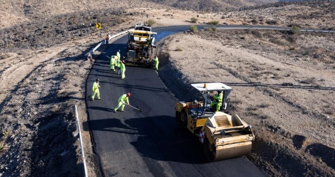 Road construction crew in high-visibility outfits laying asphalt on a winding desert road with a roller and paving machine nearby.