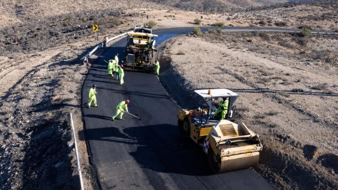 Road construction crew in high-visibility outfits laying asphalt on a winding desert road with a roller and paving machine nearby.