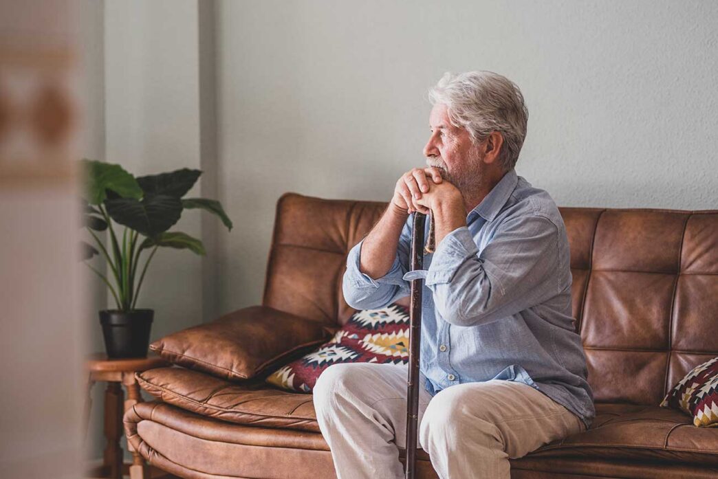 Older man with a walking cane sits on a brown leather sofa, resting chin on hands in a cozy living room.