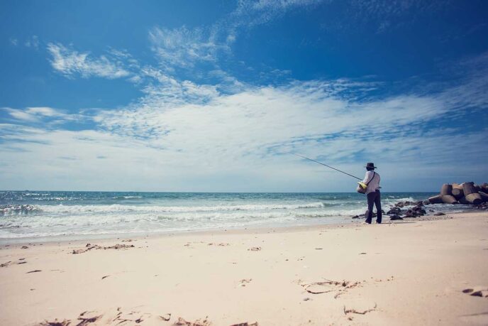 Fisherman in a hat and light shirt standing on a sandy beach, fishing into the ocean under a blue sky with scattered clouds.