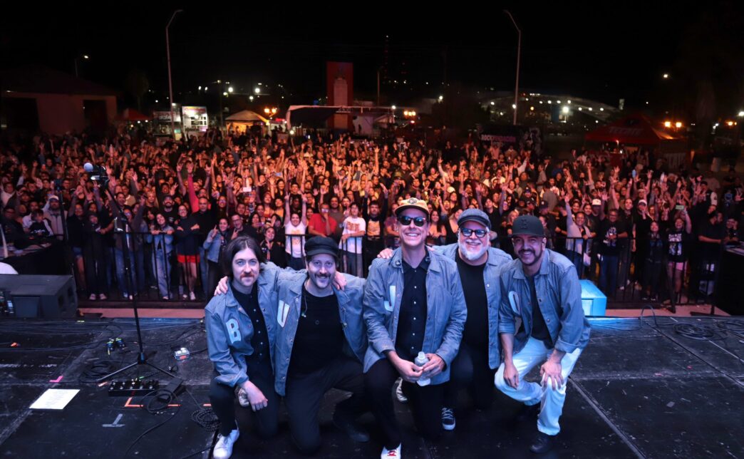 Five musicians in denim jackets kneel at the front of a stage as a large crowd cheers behind them at night.