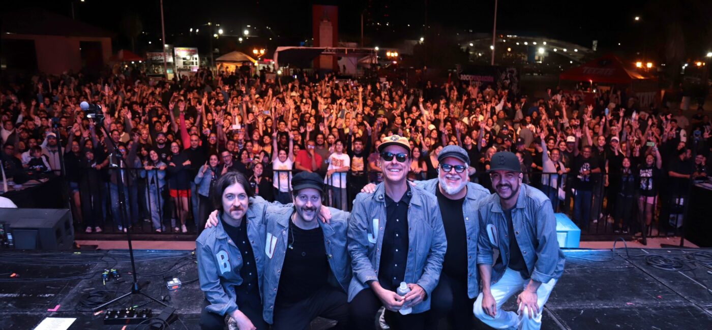 Five musicians in denim jackets kneel at the front of a stage as a large crowd cheers behind them at night.