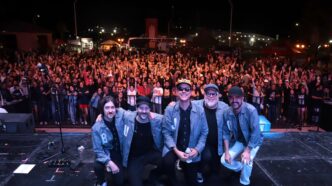 Five musicians in denim jackets kneel at the front of a stage as a large crowd cheers behind them at night.
