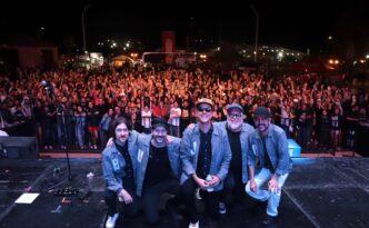 Five musicians in denim jackets kneel at the front of a stage as a large crowd cheers behind them at night.