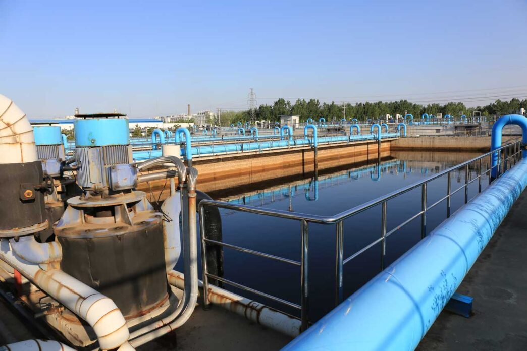 waterTreament - Gringo Gazette Industrial water treatment facility with blue pipelines and large tanks; reflective pool in the foreground and metal railing along the walkway.