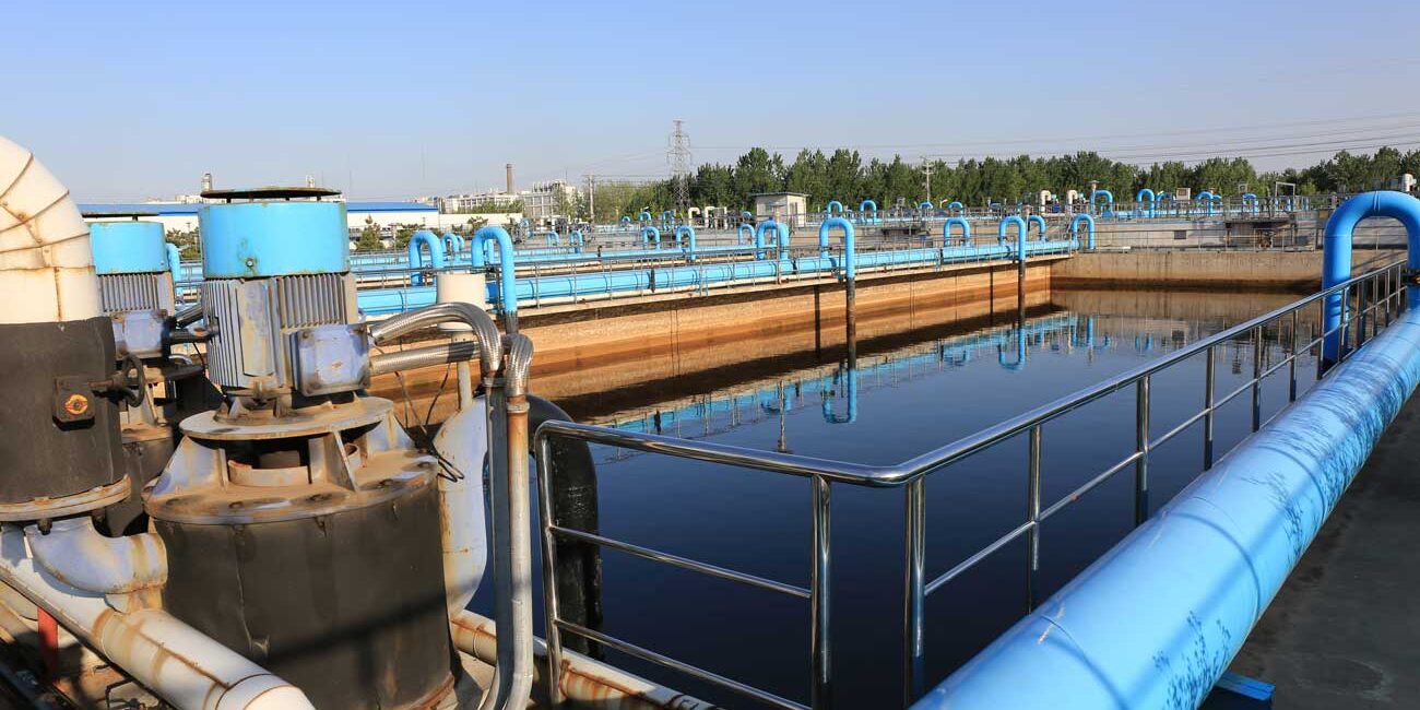 Industrial water treatment facility with blue pipelines and large tanks; reflective pool in the foreground and metal railing along the walkway.