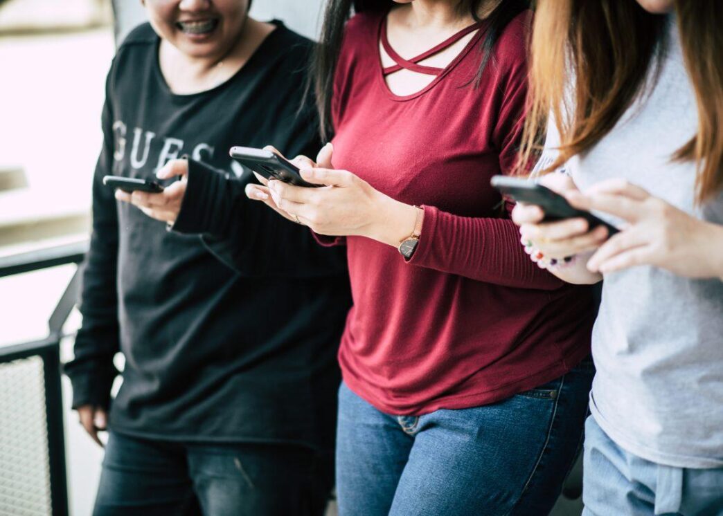 women-touch-chating-with-smart-phone-1140x815-1 - Gringo Gazette Three people standing together, each looking at a mobile phone